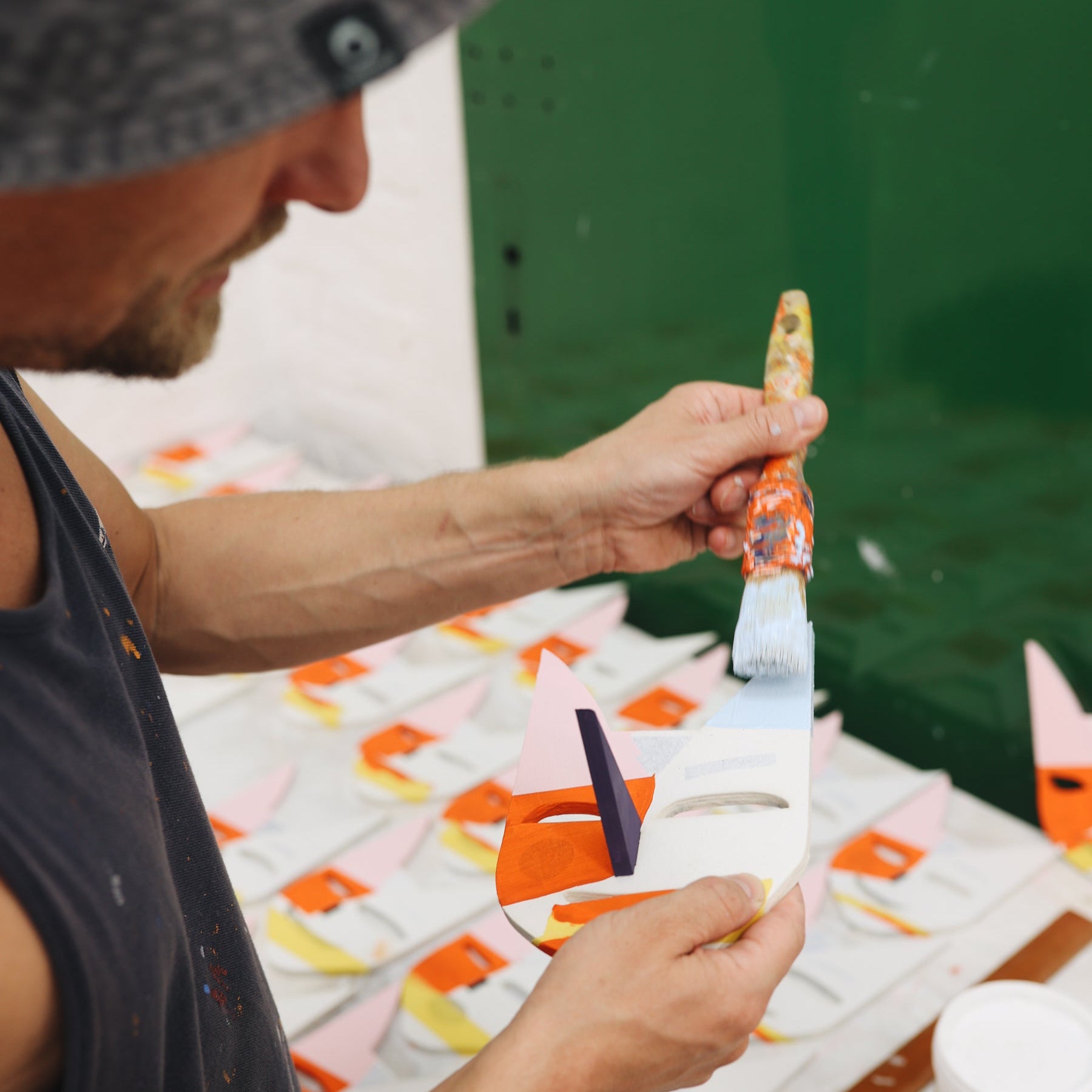 Wir schauen einem Mann über die Schulter. Er trägt ein ärmelloses Hemd und einen Sonnenhut, er steht im Atelier, hält links einen Pinsel und rechts eine Maske, die er gerade bemalt.