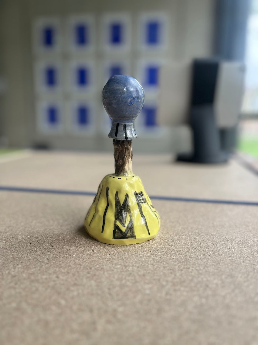 Kleinformatige Keramikskulptur „Areino“ des Künstlerduos 44flavours, hier von unten zu sehen. Es handelt sich um eine kleine Figur mit blauem, maskenartigem Kopf und langer Nase sowie einem gelben Gewand mit schwarzem Muster.