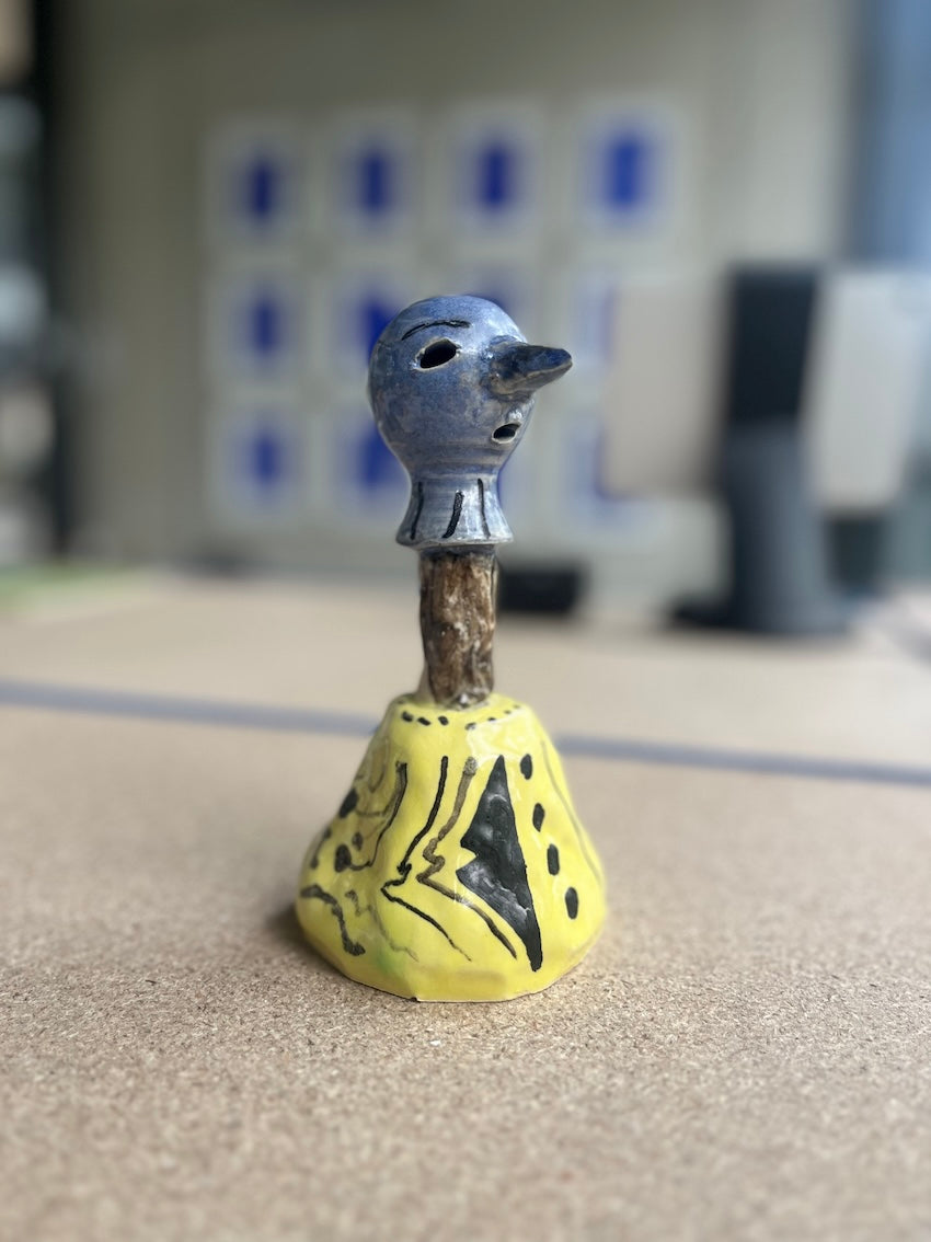 Kleinformatige Keramikskulptur „Areino“ des Künstlerduos 44flavours. Es handelt sich um eine kleine Figur mit blauem, maskenartigem Kopf und langer Nase sowie einem gelben Gewand mit schwarzem Muster.