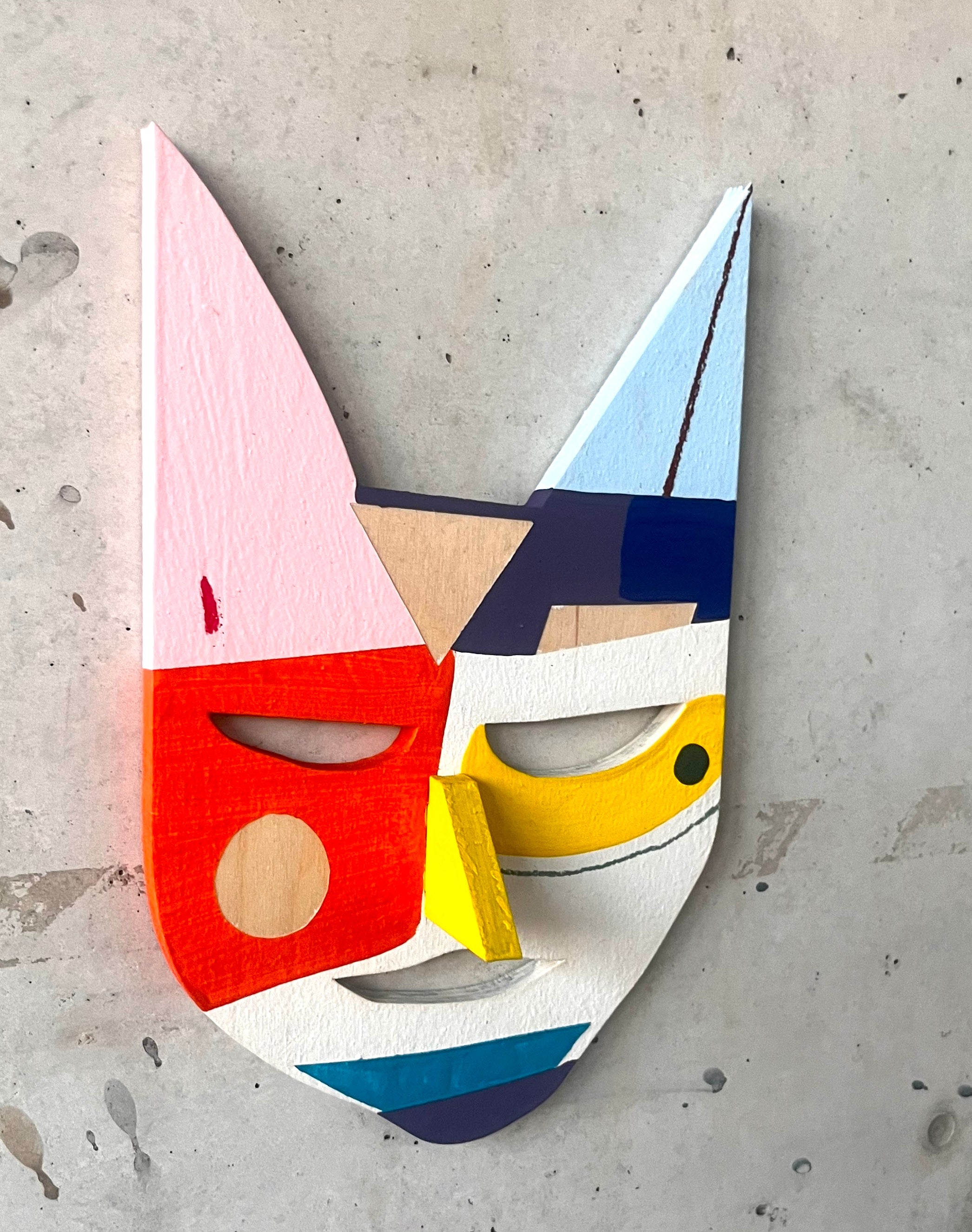 An einer grauen Wand hängt eine bunt bemalte Holzmaske 