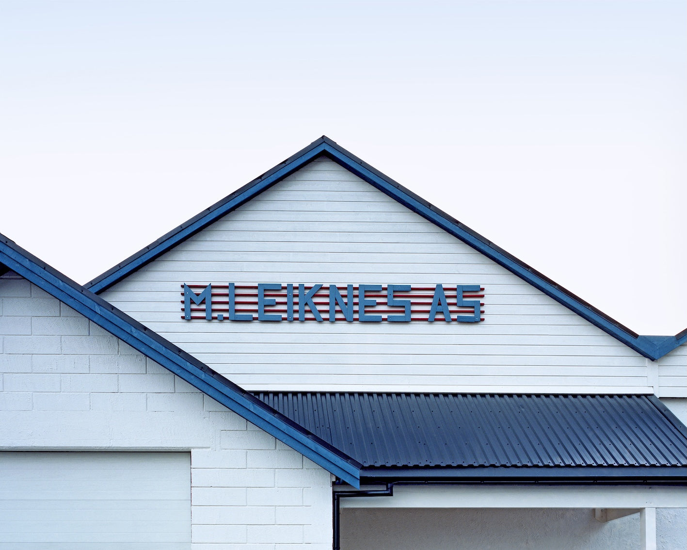 In einer scheinbar ruhigen abendlichen Stimmung sieht man das Dach und Vordach eines Holzhauses, das hellgrau und blau gestrichen ist. Auf dem Giebel des Daches ist der Firmenname Leiknes AS zu lesen