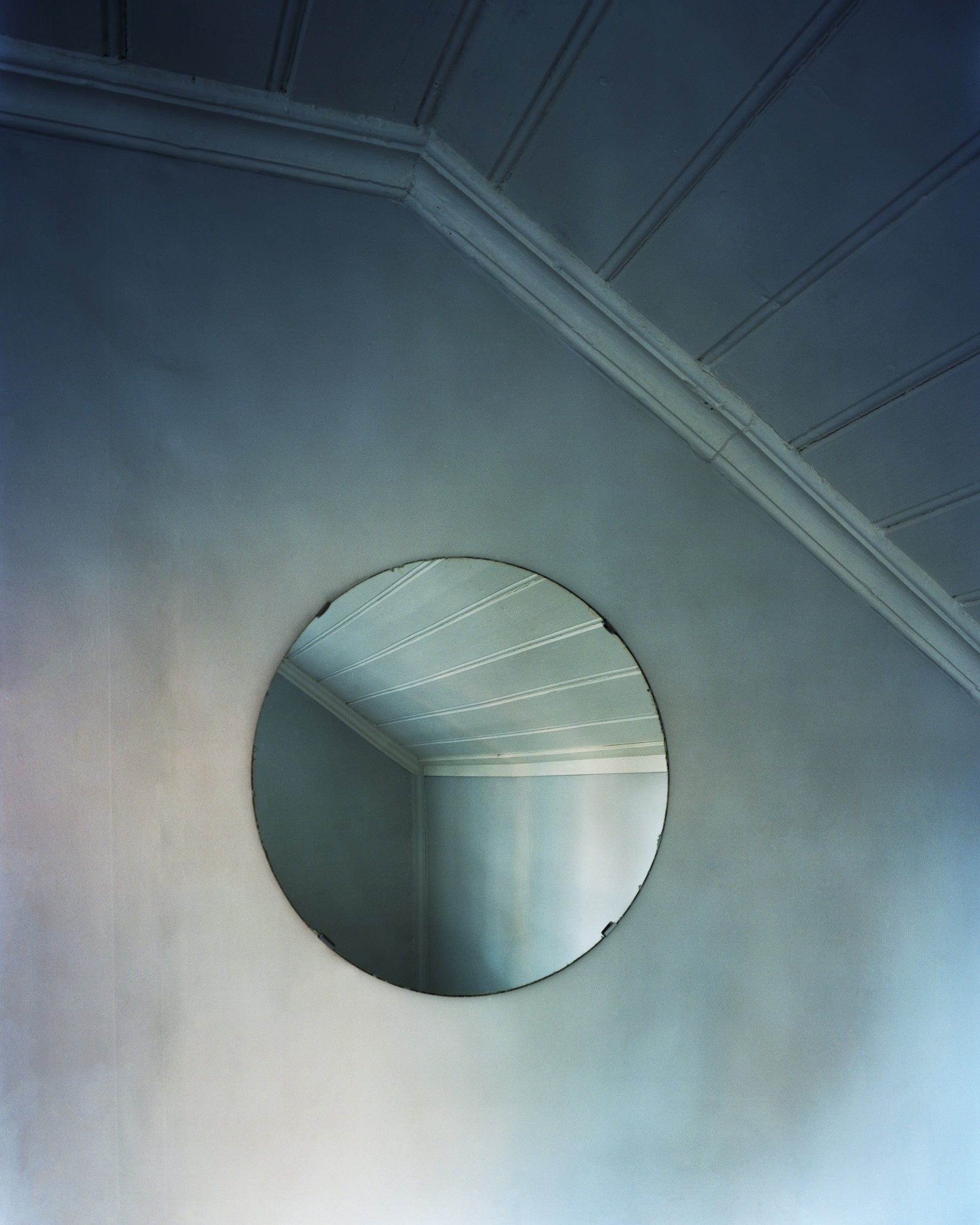 An der Wand im Treppenaufgang eines offenbar alten Hauses hängt ein runder Spiegel, in dem man die gegenüberliegende Zimmerecke sieht. Wände und Decke sind in einem hellen Grau gestrichen, von der linken Seite fällt Tageslicht ein