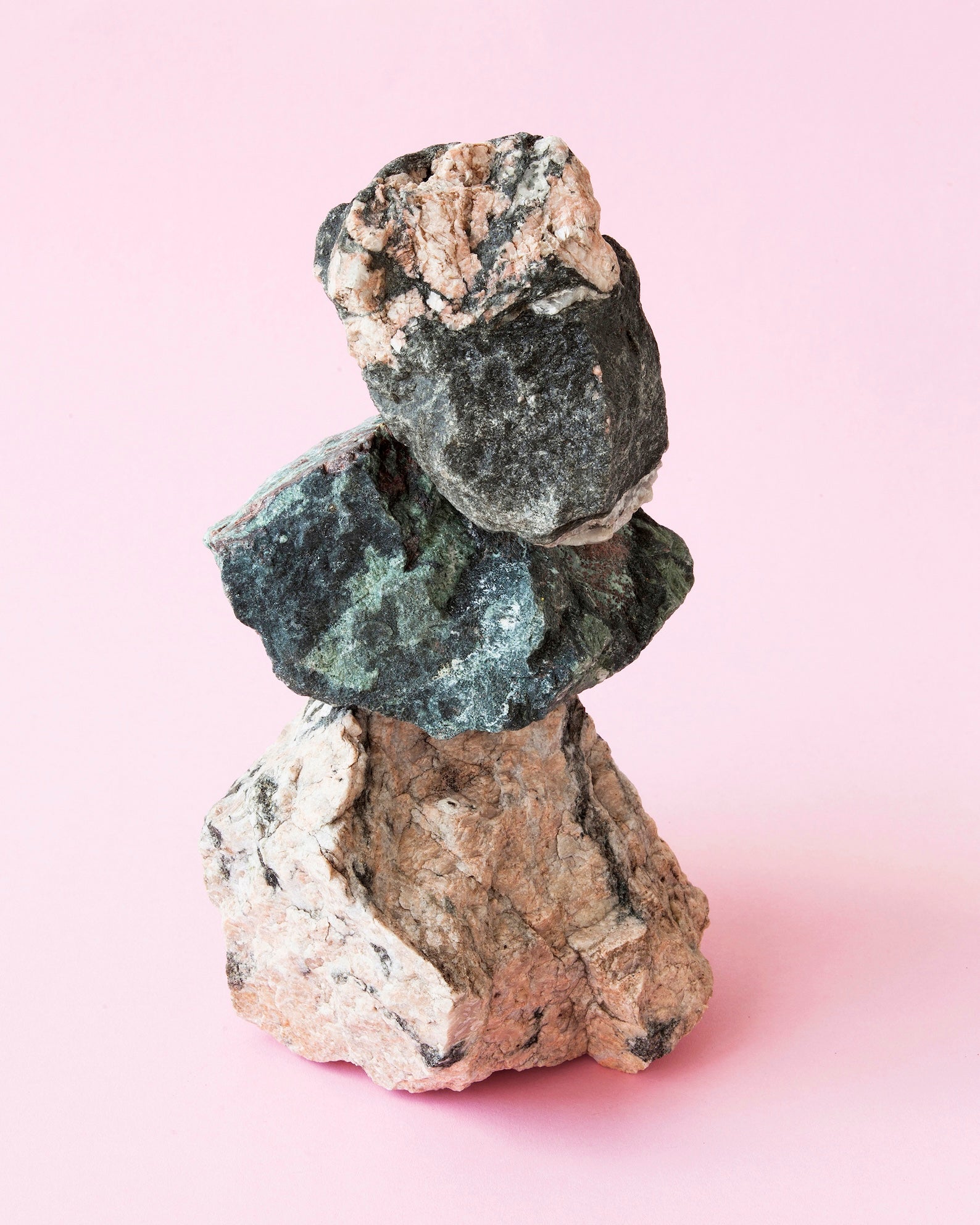 Vor einem einfarbigen rosa Hintergrund balanciert ein kleiner Haufen aus drei beige-grauen Steinen. Die Steine sind unbehauen und grob, und es sieht so aus als könnte der obere gleich runterfallen