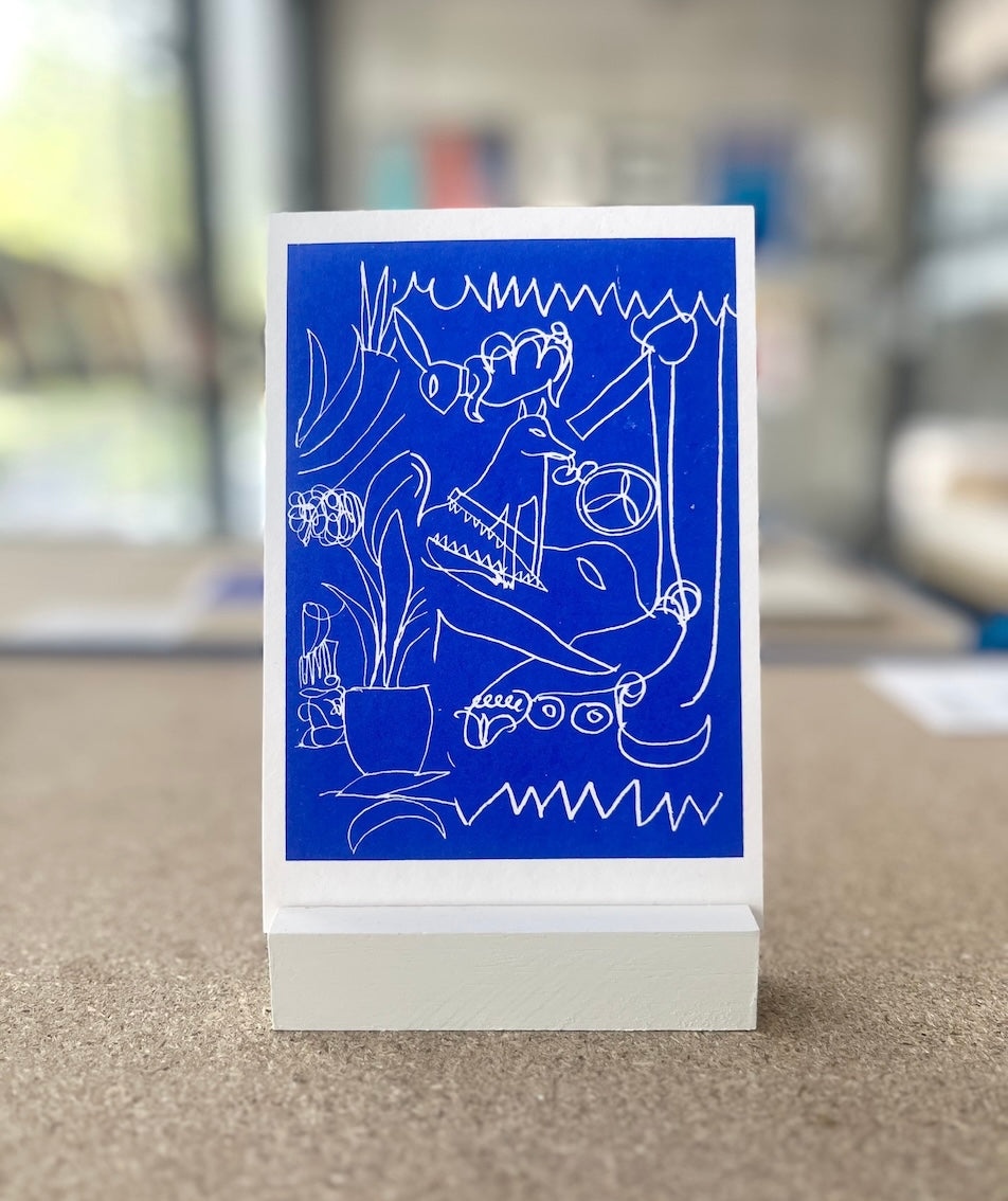 Ein kleinformatiger Druck wird in einem kleinen Holzständer präsentiert. Der Druck ist tiefblau und hat weiße Linienzeichnungen: Ein Krokodil, ein Hund, Muster und Pflanzen. Der Raum im Hintergrund ist verschwommen. 