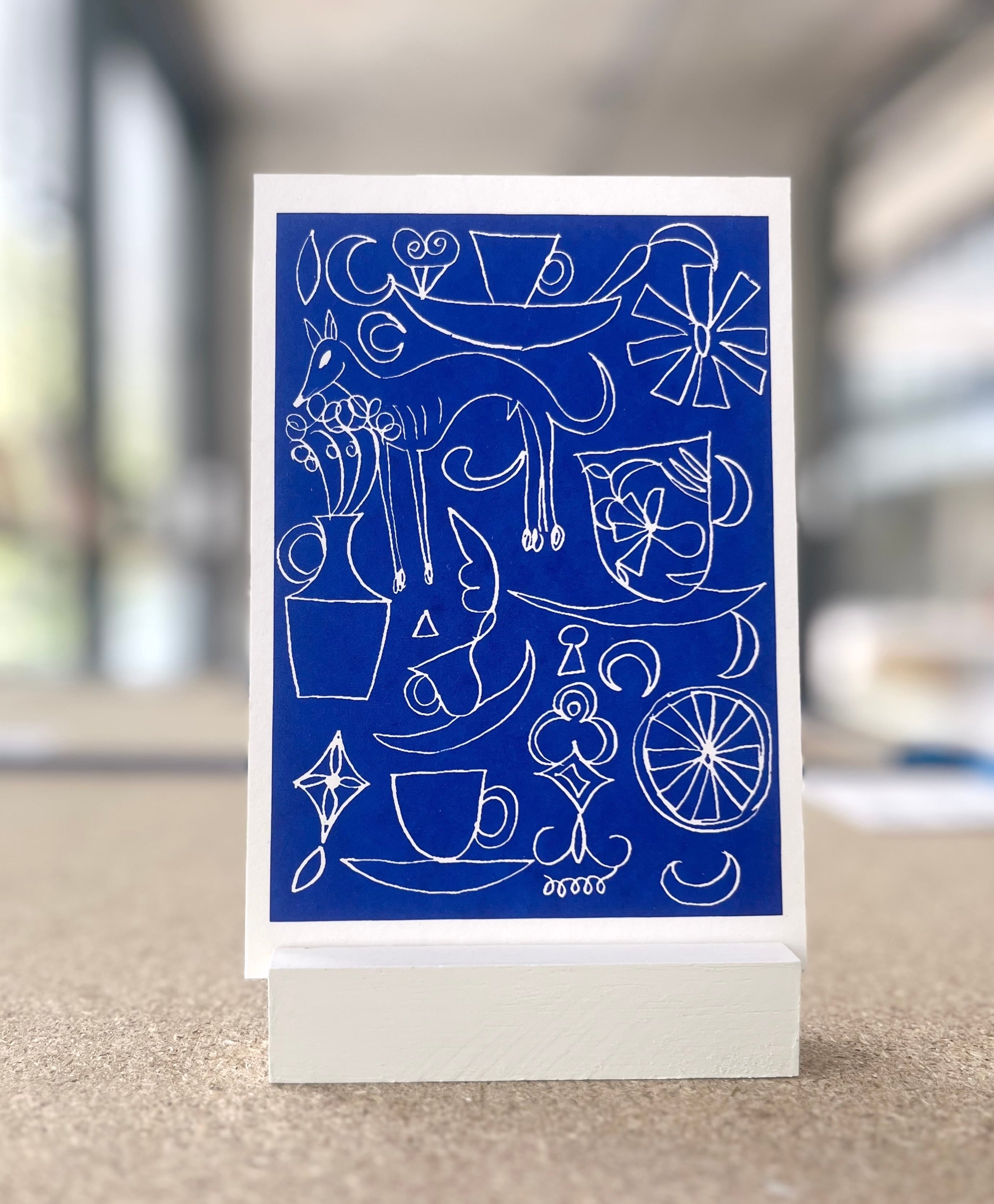 Ein kleinformatiger Druck wird in einem kleinen Holzständer präsentiert. Der Druck ist tiefblau und hat weiße Linienzeichnungen: Pflanzenmotive, einen schlanken Hund, aber auch eine Tasse. Der Raum im Hintergrund ist verschwommen. 