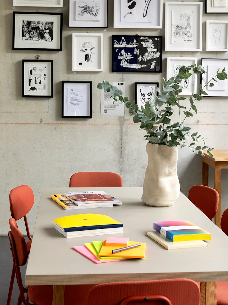 Blick in den Raum von Zelt Berlin vorbereitet für einen Workshop. Auf dem Tisch liegen Arbeitsmaterialien und ein Blumenstrauß. Im Hintergrund an der Wand hängen viele gerahmte Originalzeichnungen.  