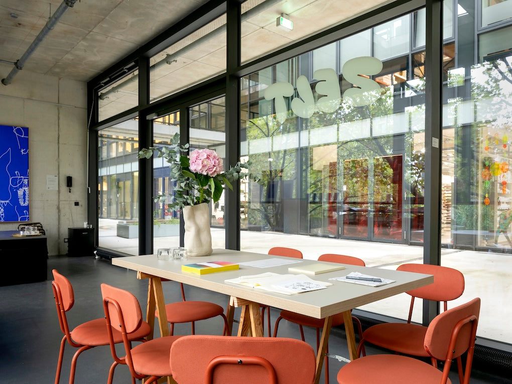 Ein Blick in den Raum von Zelt Berlin als Workshop-Space. Mit einem großen Tisch und Stühlen, Arbeitsmaterialien und einem Blumenstrauß auf dem Tisch. Der Blick geht durch das große Fenster auf die Terrasse. Im Hintergrund ist ein riesiger blauer Siebdruck von Simon Blume zu sehen. 