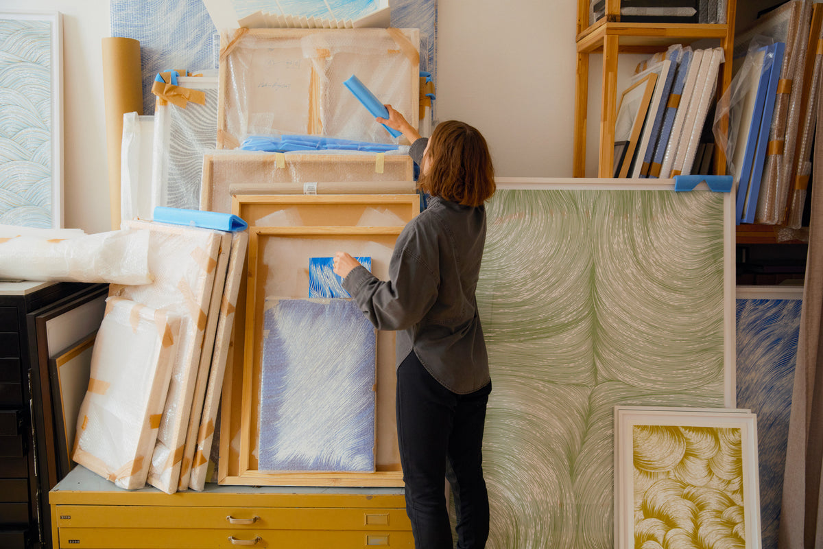 Die Künstlerin Bettina Krieg in ihrem Atelier, viele eingepackte Bilder, einige Arbeiten sind gerahmt aufgestellt und zeigen ihre typischen Linienformationen. 